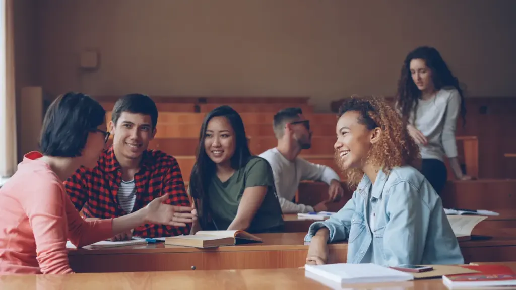 Diverse students laughing and talking together at desks in a university lecture hall.